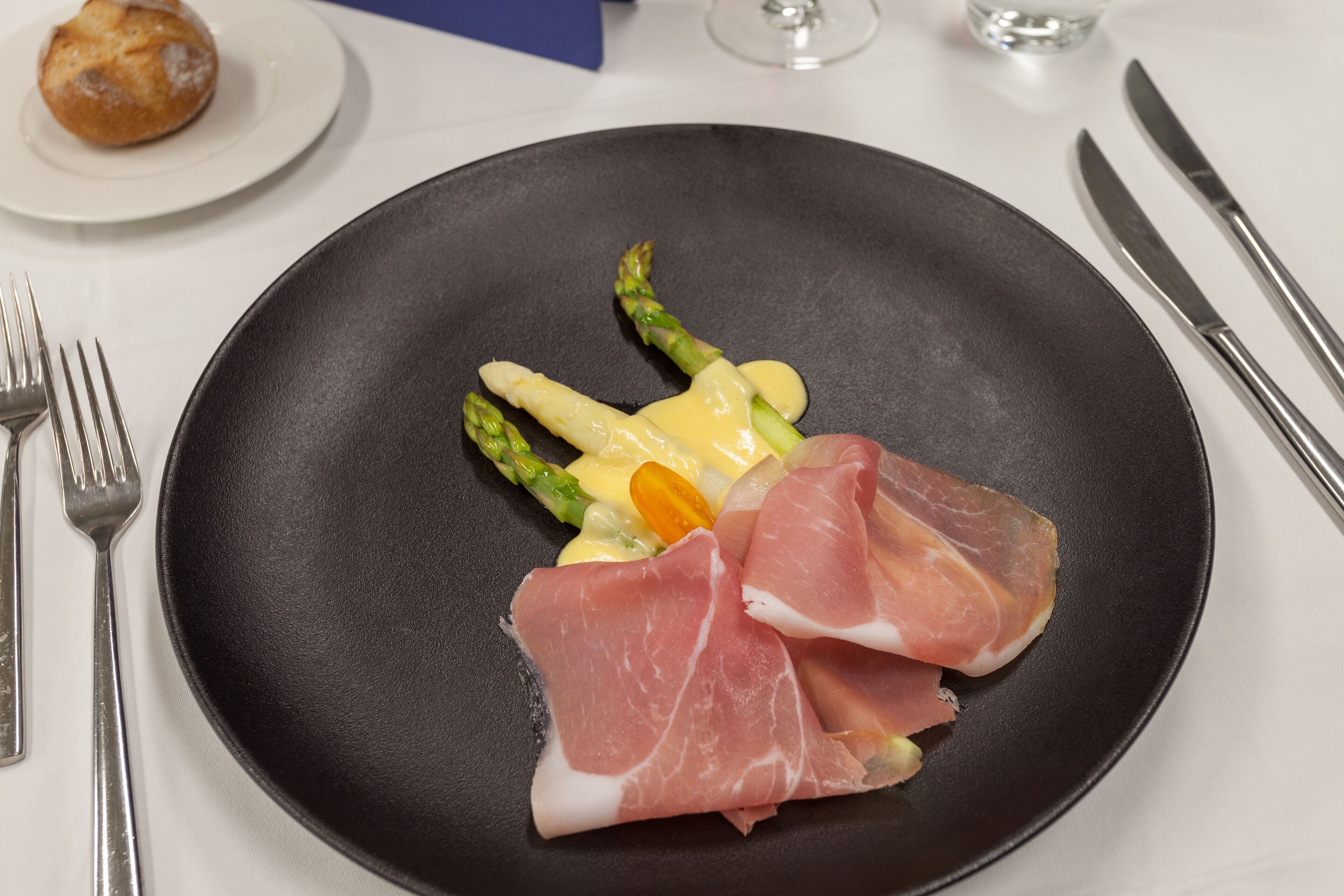 A black plate with asparagus and prosciutto on a white tablecloth.