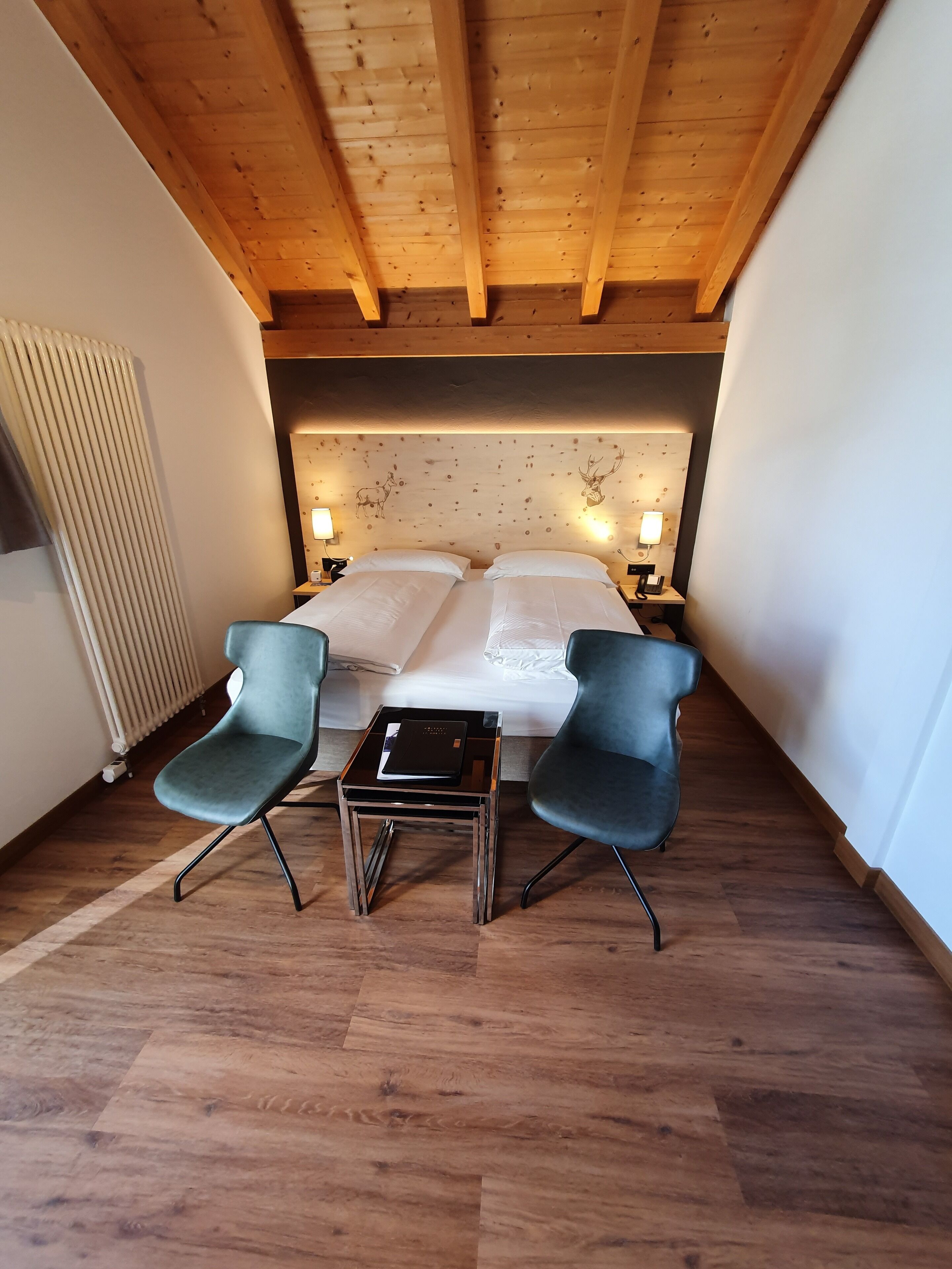 Un dormitorio con techo de madera, dos camas, dos sillas, una mesita, lámparas de noche y un radiador.