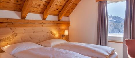 Techo de madera con vigas a la vista, cama con ropa blanca, lámpara de pared y ventana con cortinas.