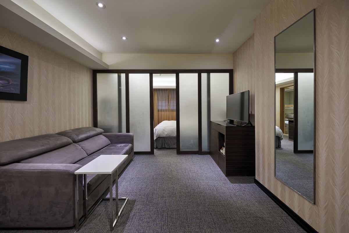 A modern hotel room with a sofa, a flat-screen TV, a large mirror, and a sliding door leading to a bedroom.