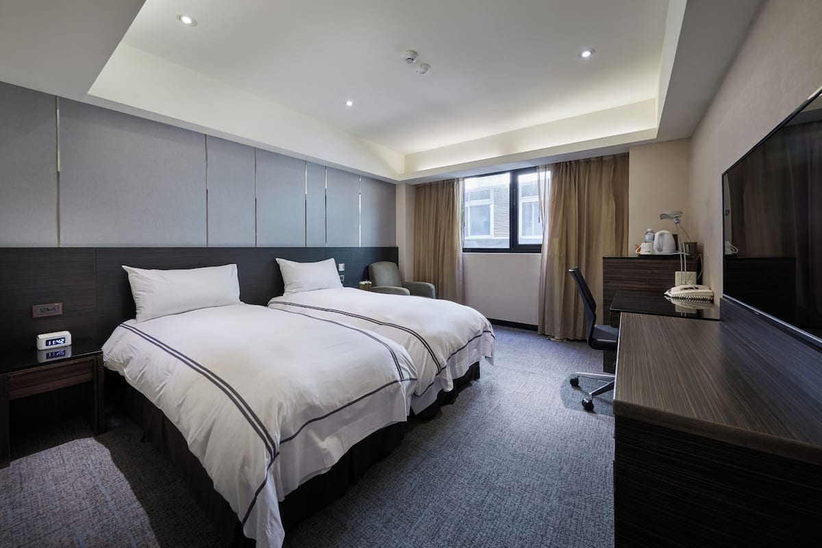 A hotel room with two beds, a desk, a chair, a TV, and a window with curtains.
