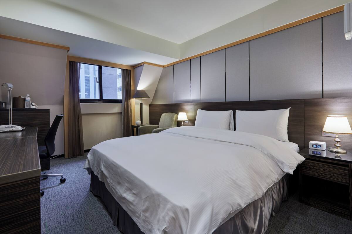 A hotel room with a large bed, a desk with a lamp, a chair, a nightstand with a clock, and a window with curtains.