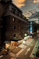 Un hotel de varios pisos con un diseño arquitectónico moderno, destacando una entrada prominente, una escalera visible y una vista clara de la calle con flechas direccionales.