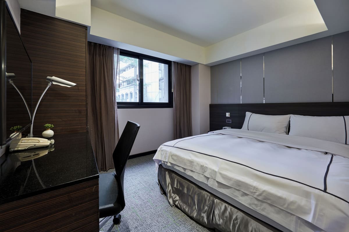 A hotel room with a bed, a desk with a lamp, a chair, and a window with curtains.