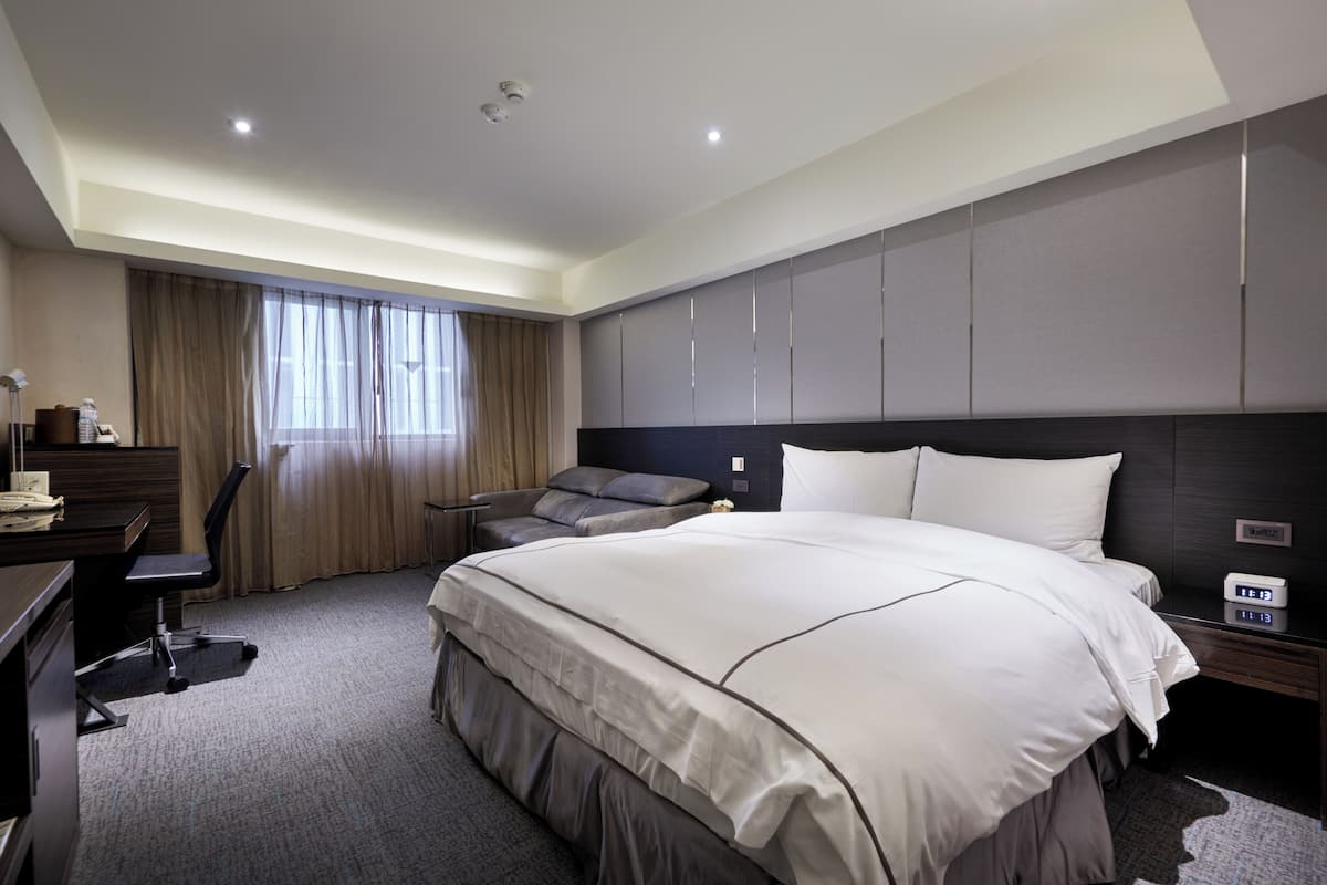 A modern hotel room with a large bed, a desk, a chair, and a nightstand with a digital clock.