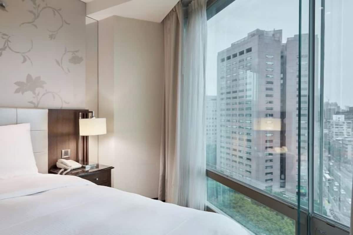 Habitación de hotel con cama, mesita de noche con lámpara y vistas a edificios altos a través de amplias ventanas.