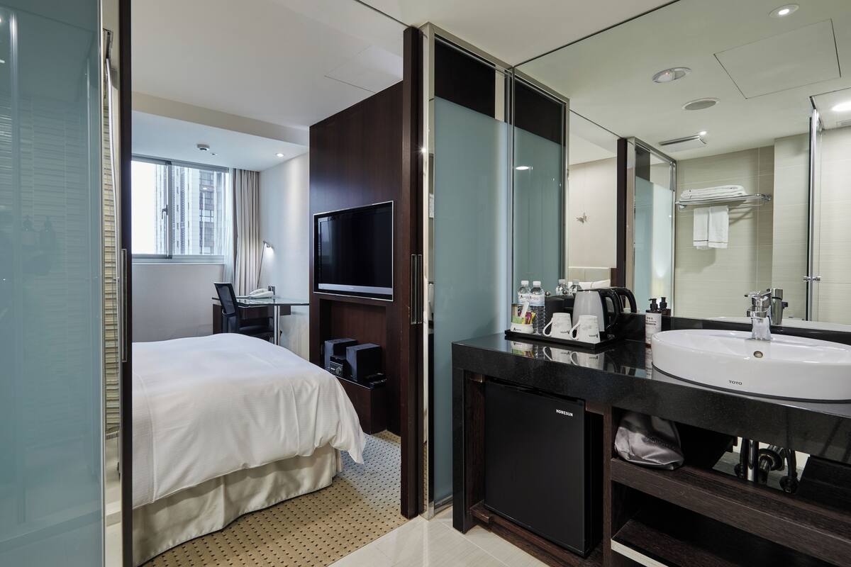 Habitación de hotel con cama, televisión, minibar, espejo grande y baño con lavamanos y ducha.