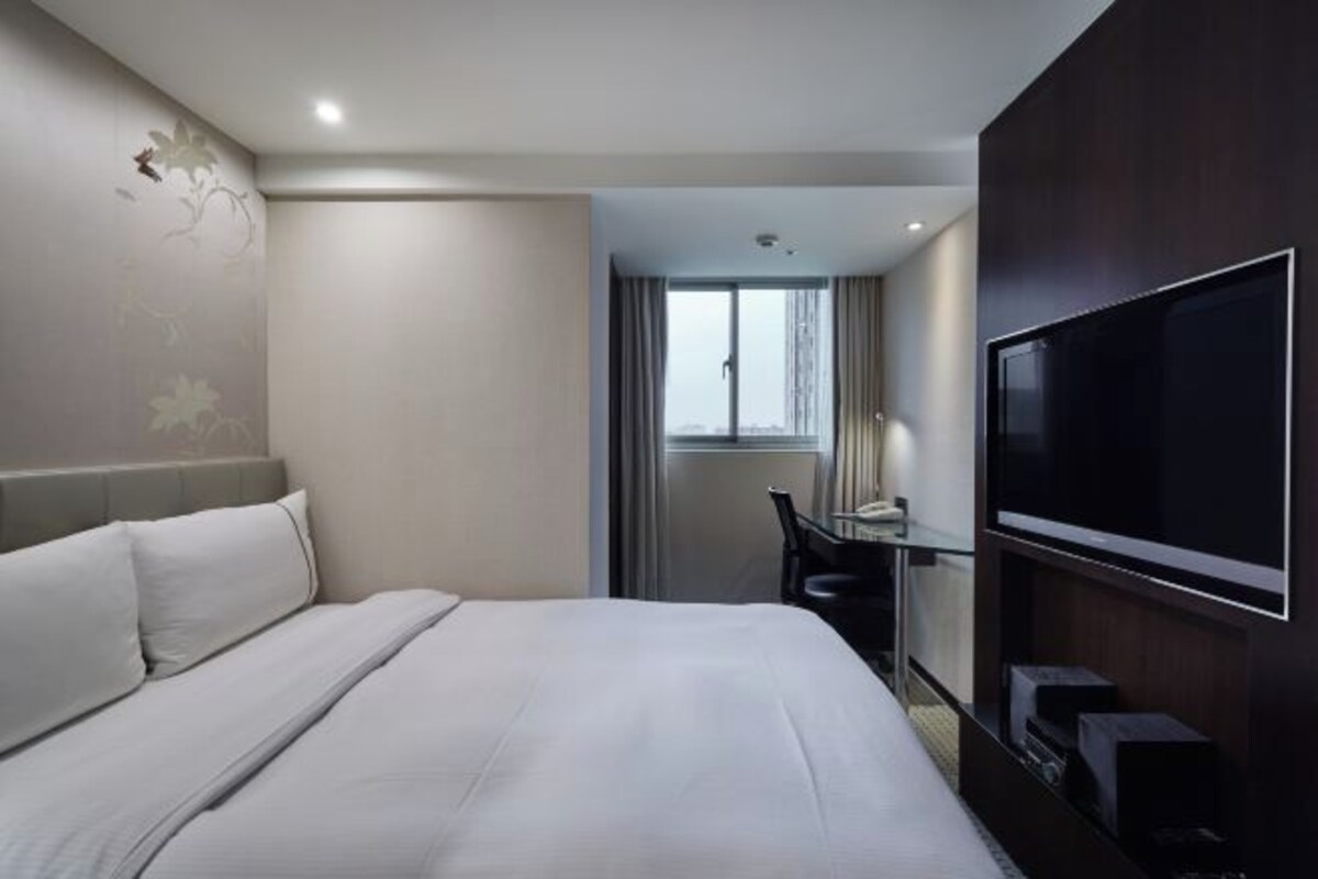 A hotel room with a bed, a desk with a chair, a TV, and a window.