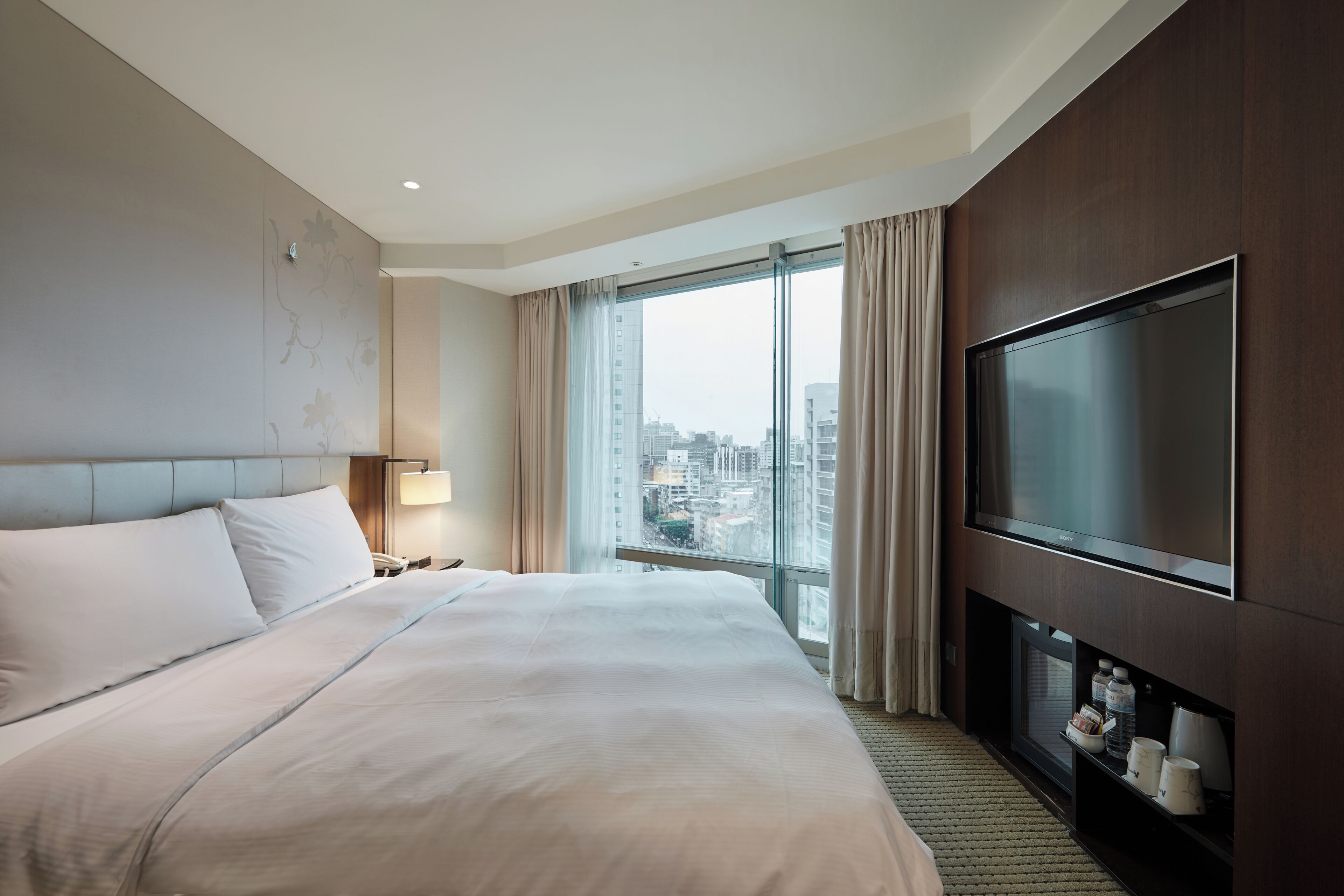 A hotel room with a large bed, a television, a nightstand, and a view of the city through large windows.