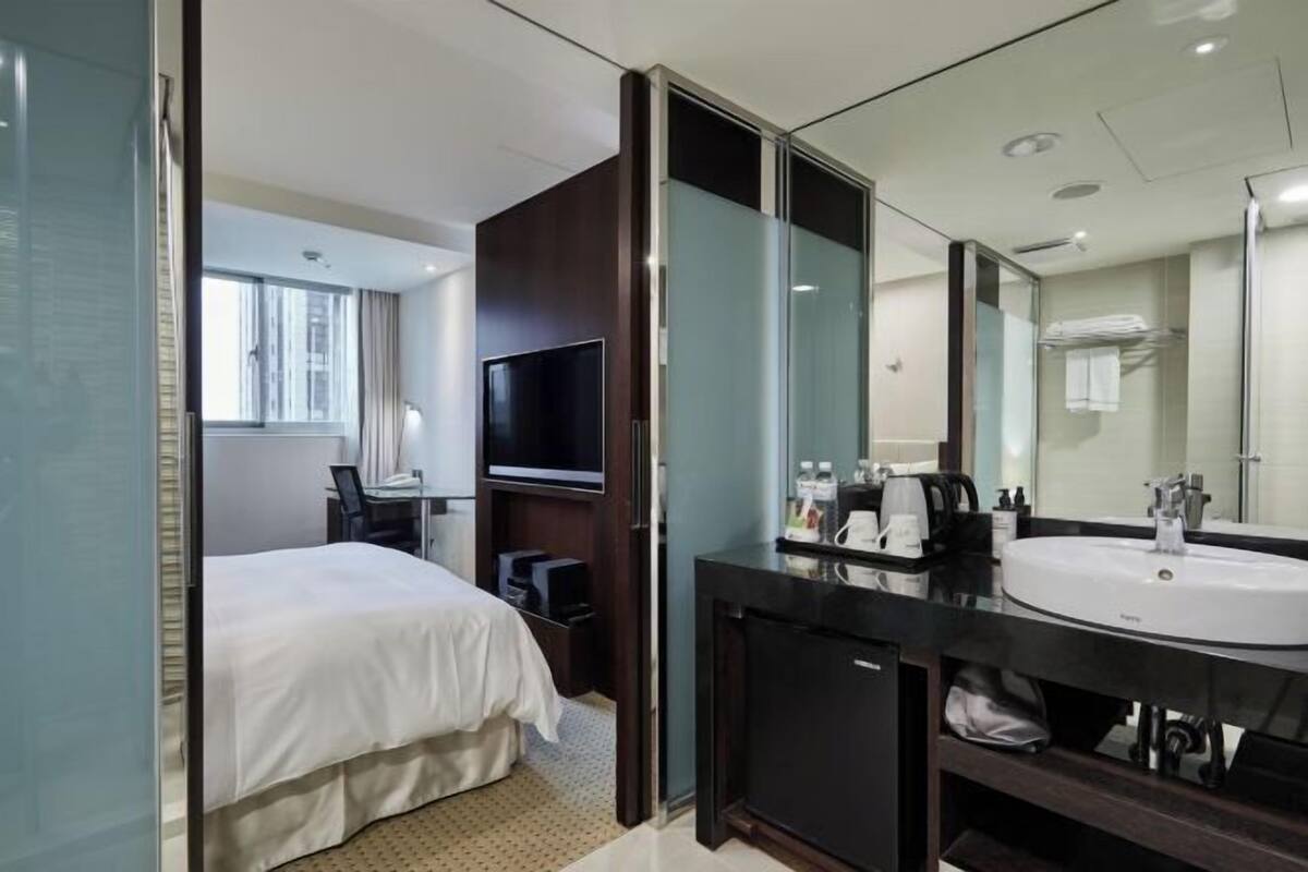Habitación de hotel con cama, televisión, escritorio, espejo grande y baño con lavamanos y ducha.