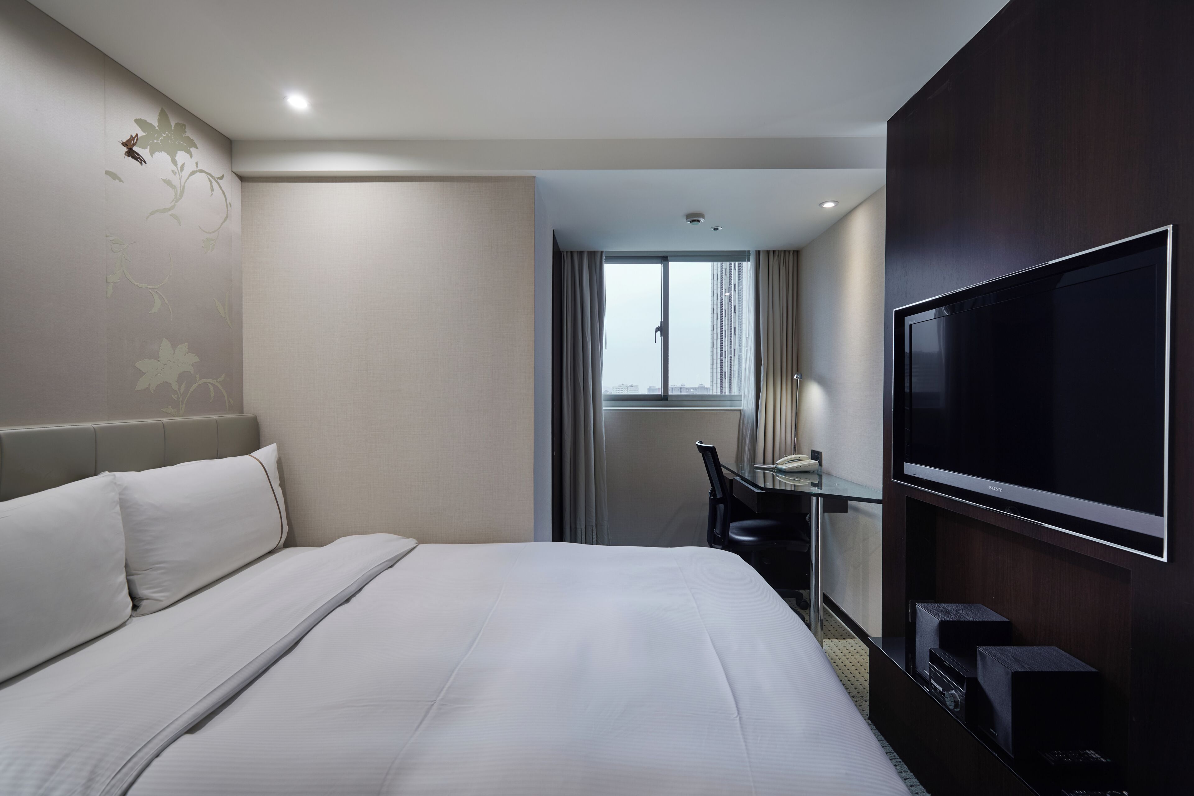 A hotel room with a large bed, a flat-screen TV, a desk with a chair, and a window with curtains.