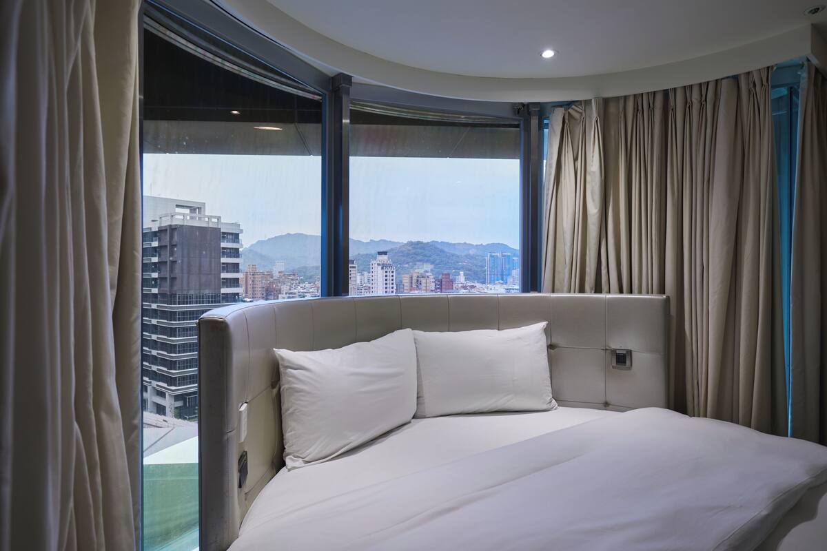 Habitación de hotel con una cama grande, cabecera y vistas a un paisaje urbano a través de amplias ventanas.