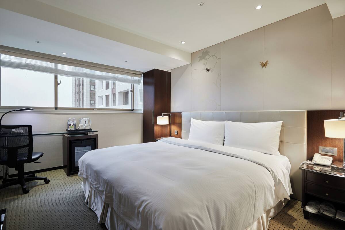 Habitación de hotel con una cama grande, un escritorio con silla, un refrigerador pequeño y una ventana con persianas.