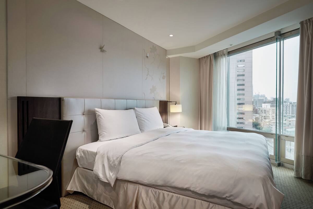 Habitación de hotel con cama, una silla, una mesa y un ventanal con vistas a la ciudad.