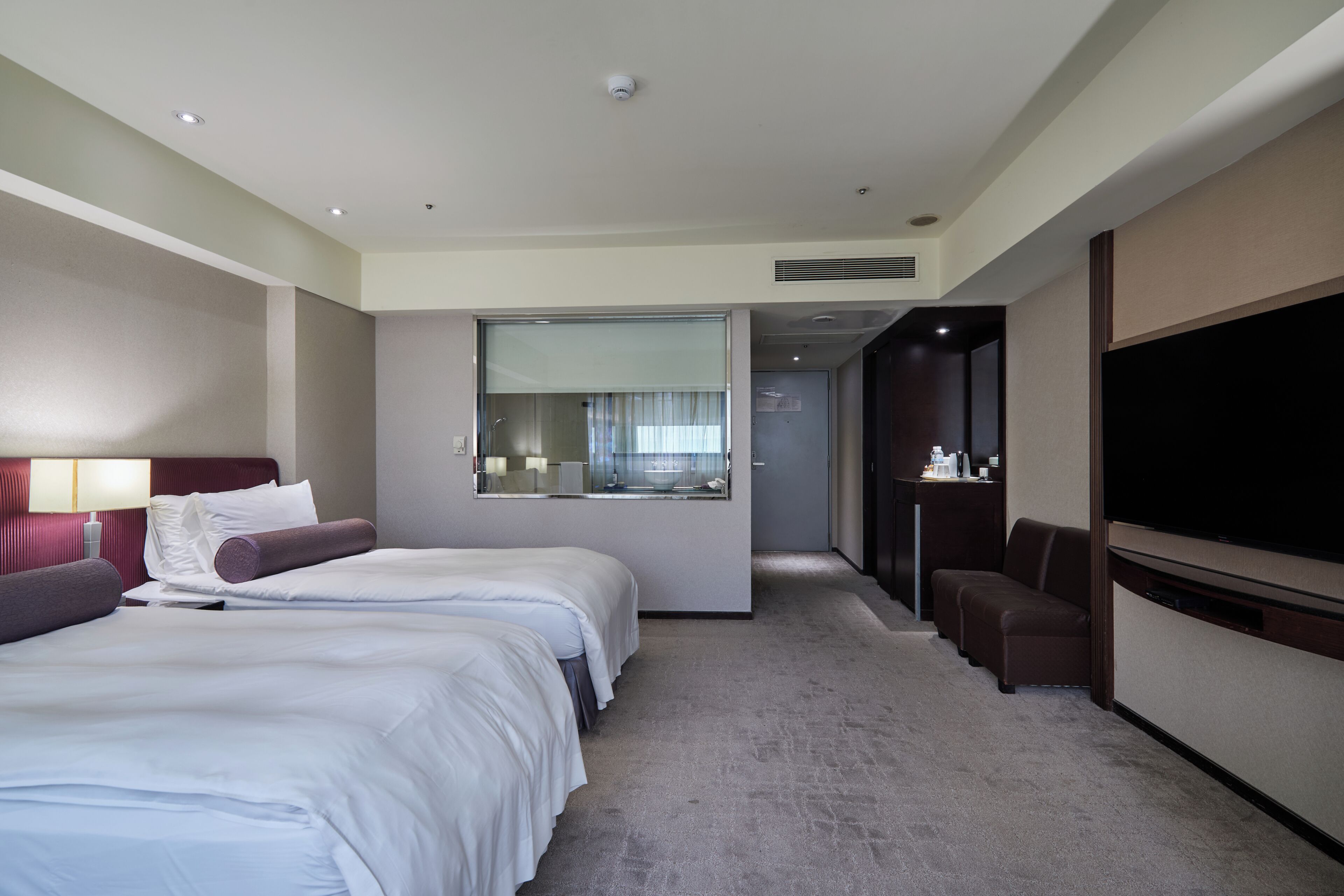 A hotel room with a large bed, a TV, a seating area, and a bathroom visible through a glass door.