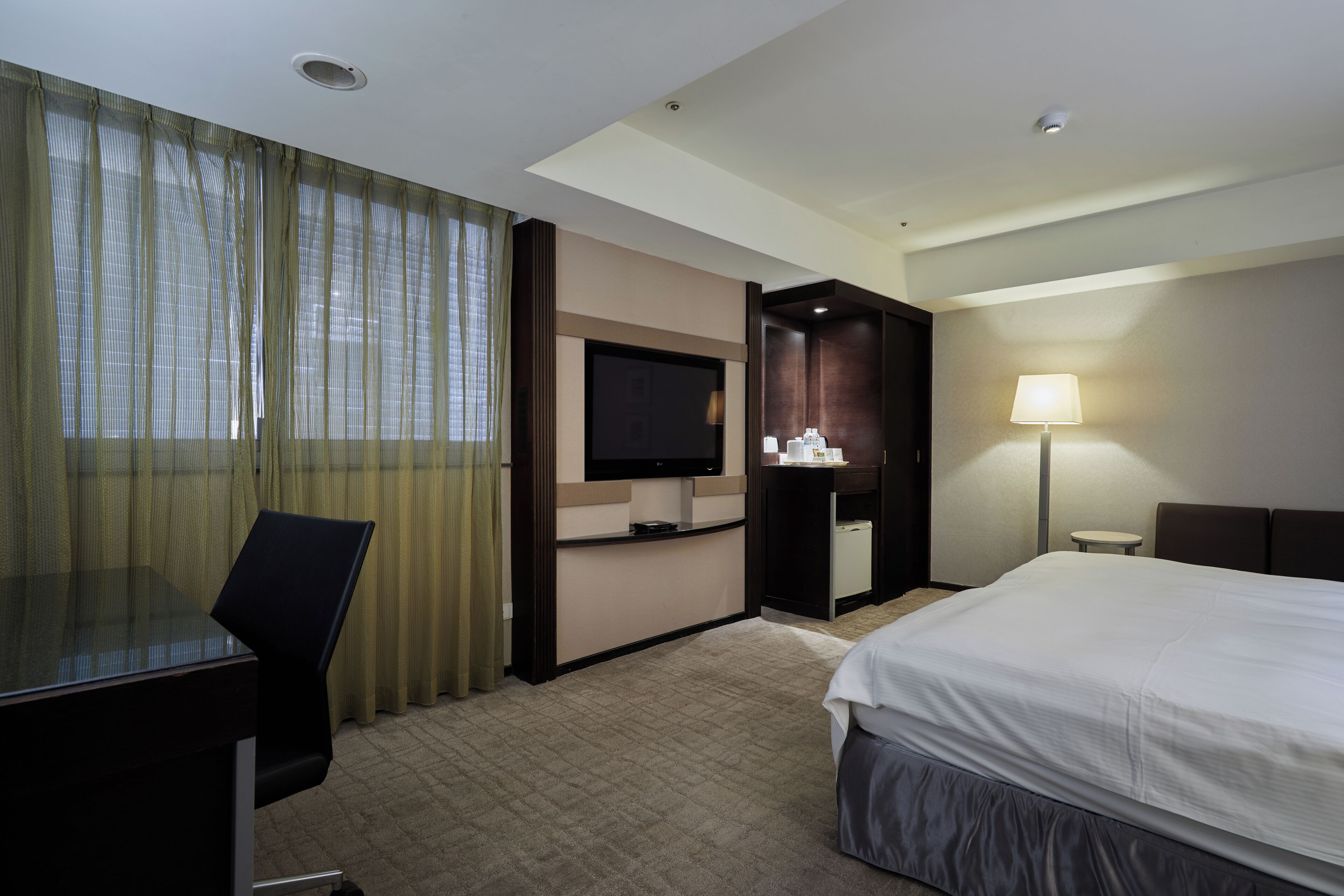 Een hotelkamer met een bed, een bureau, een televisie, een lamp en een raam met gordijnen.