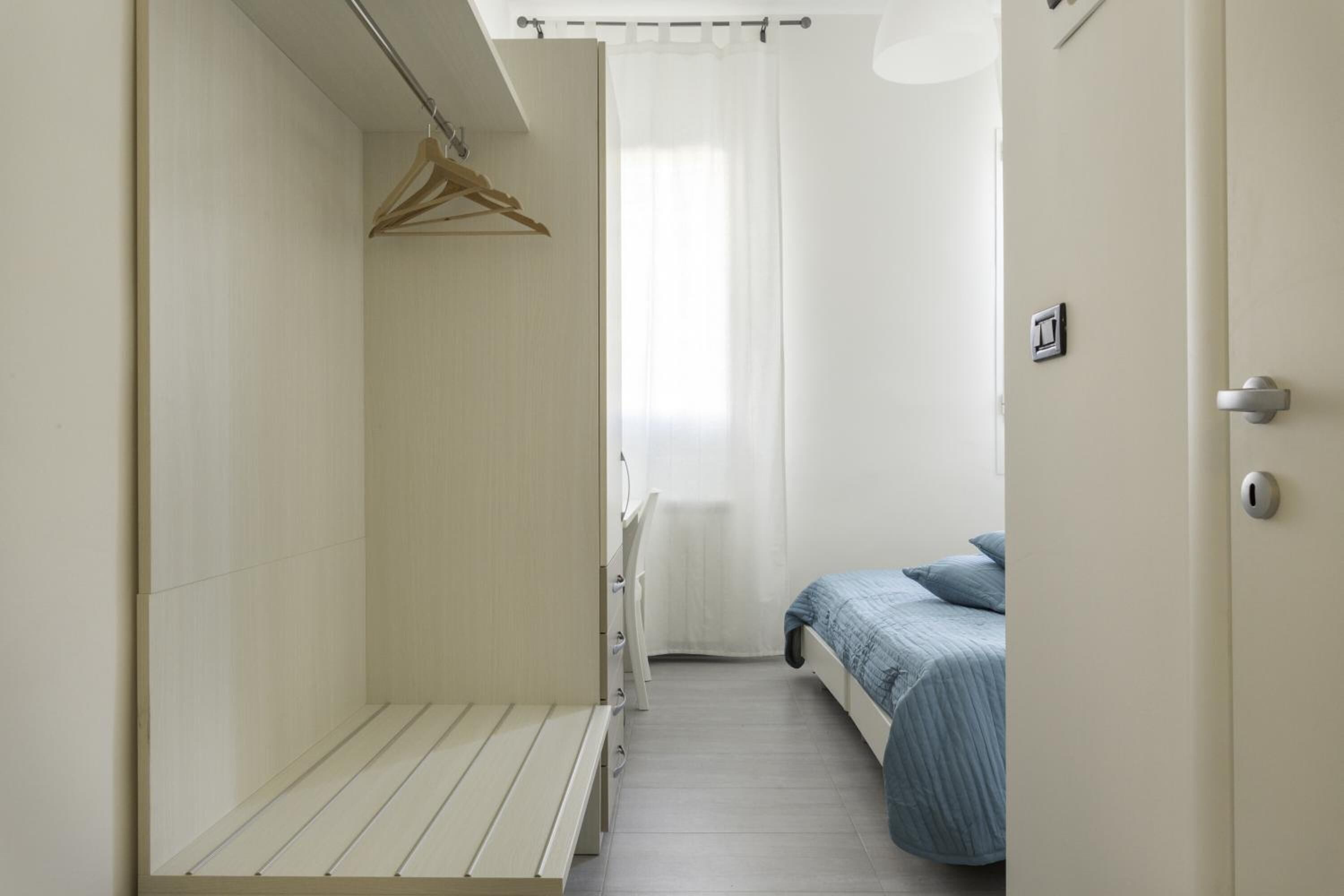 A narrow room with a bed, a bench, and a hanging clothes rack.