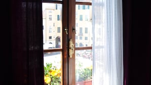 A room with a window overlooking a cityscape, featuring a wooden door and a view of buildings and flowers outside. - At Home Bed and Breakfast (Lucca)