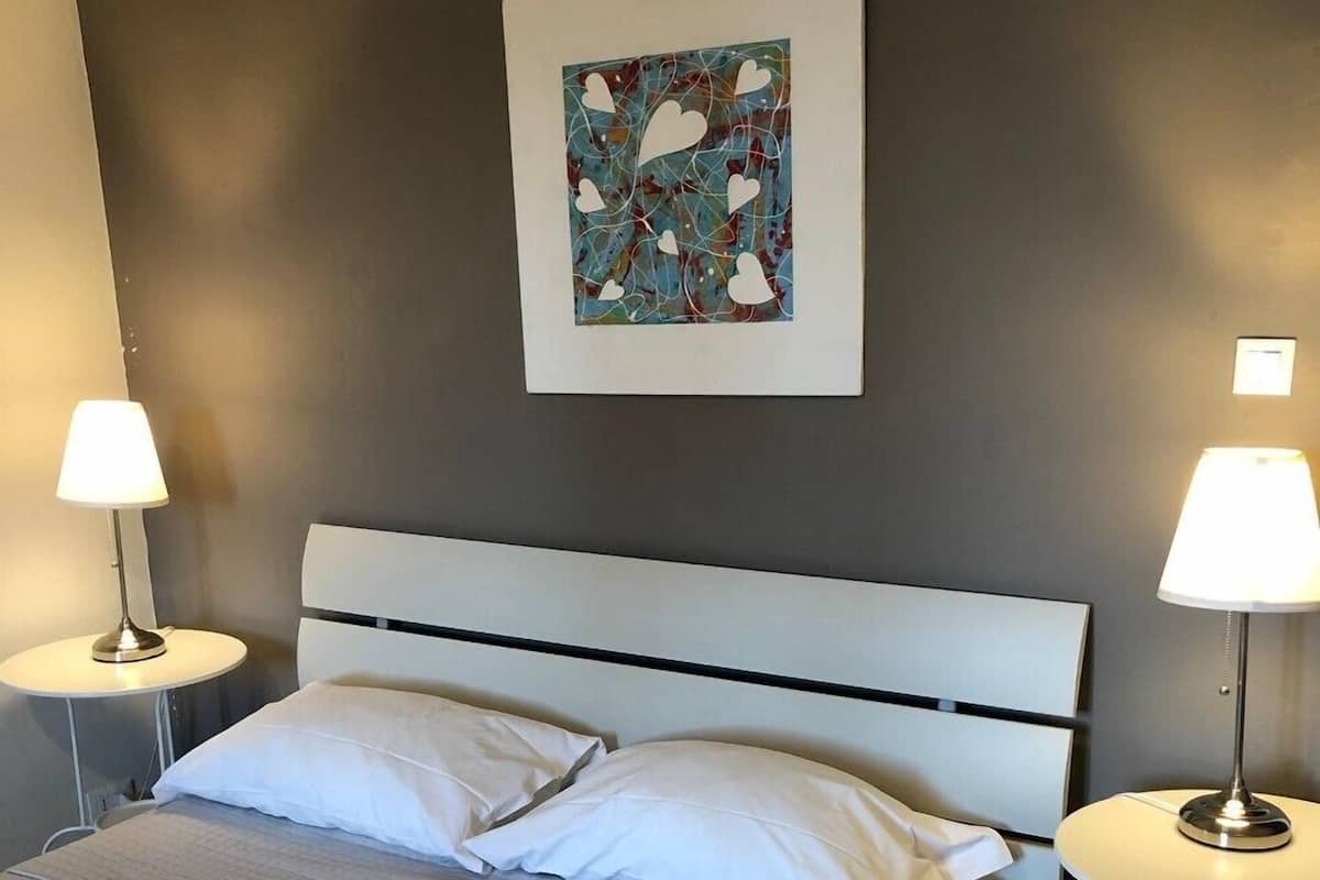 Una cama bien hecha con sábanas blancas, dos lámparas de noche y una obra de arte abstracta colgada en la pared.
