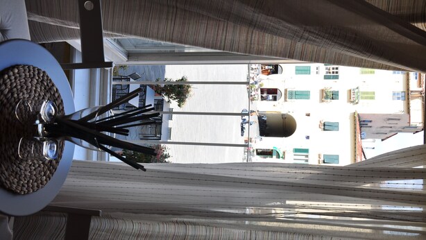 A view through a window with striped blinds, a white table with a decorative plate, and a street scene with buildings and parked cars. - At Home Bed and Breakfast (Lucca)