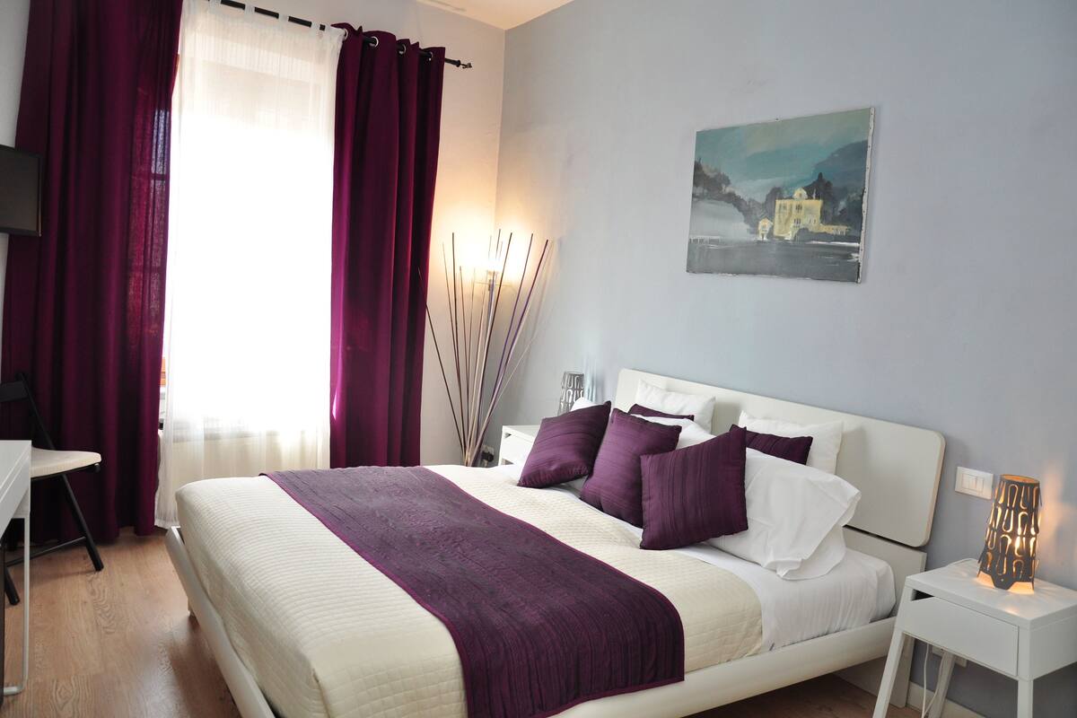 Un dormitorio ordenado con cama, mesita de noche, silla y un cuadro en la pared.