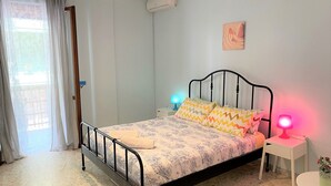 Un dormitorio con cama, dos lámparas de noche, una mesita, una planta en maceta y una ventana con cortinas.