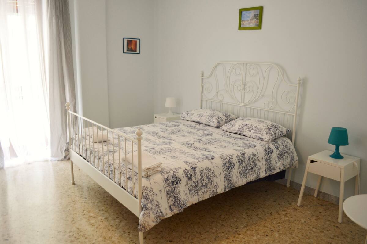 Un dormitorio con una estructura de cama de metal blanco, una colcha estampada, una mesita de noche con lámpara y una pequeña imagen en la pared.