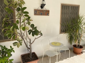 A small patio area with a potted tree, two white metal chairs, a small table with a yellow placemat, and a wooden sign with the name 'Olive Tree'.