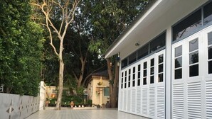 A covered outdoor area with patterned flooring, a white building with multiple windows, and trees in the background. - Baan Suksabai Hua Hin (Hua Hin)
