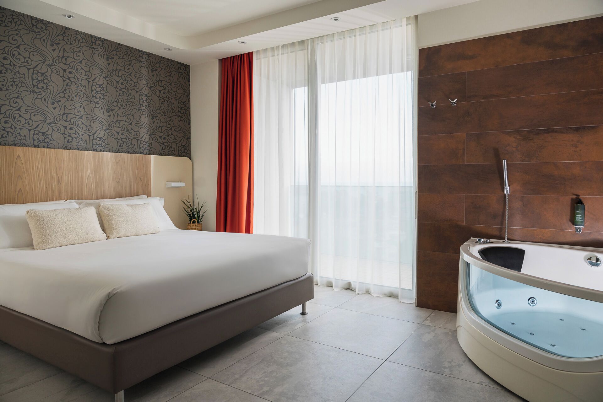 Una camera d'albergo moderna con un letto grande, una vasca da bagno vetrata e vista sulla città attraverso ampie finestre.