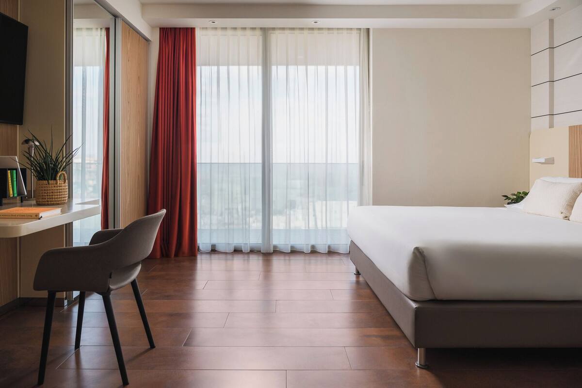 Une chambre d’hôtel avec un lit, un bureau et une chaise, une fenêtre avec des rideaux et une plante sur le bureau.