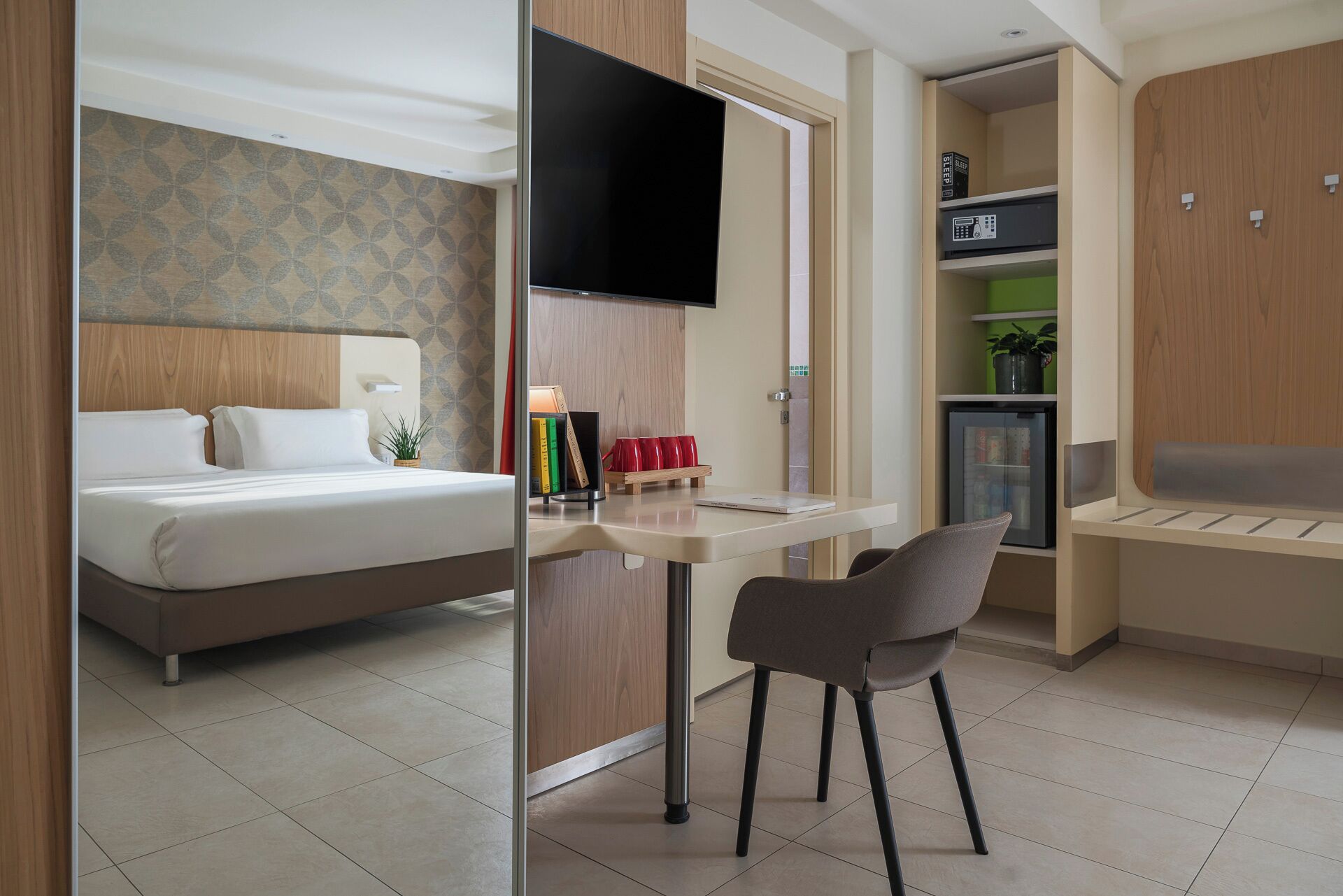 Una moderna camera d'albergo con un letto, una scrivania con una sedia, una TV, un mini-frigorifero e una piccola pianta.