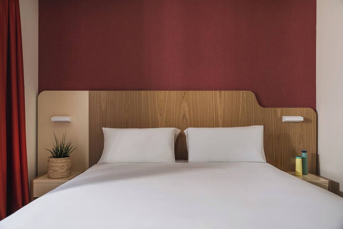 Une chambre d’hôtel avec une tête de lit en bois, deux oreillers blancs, un couvre-lit blanc, un rideau rouge, une petite plante et une table de chevet avec une bouteille.