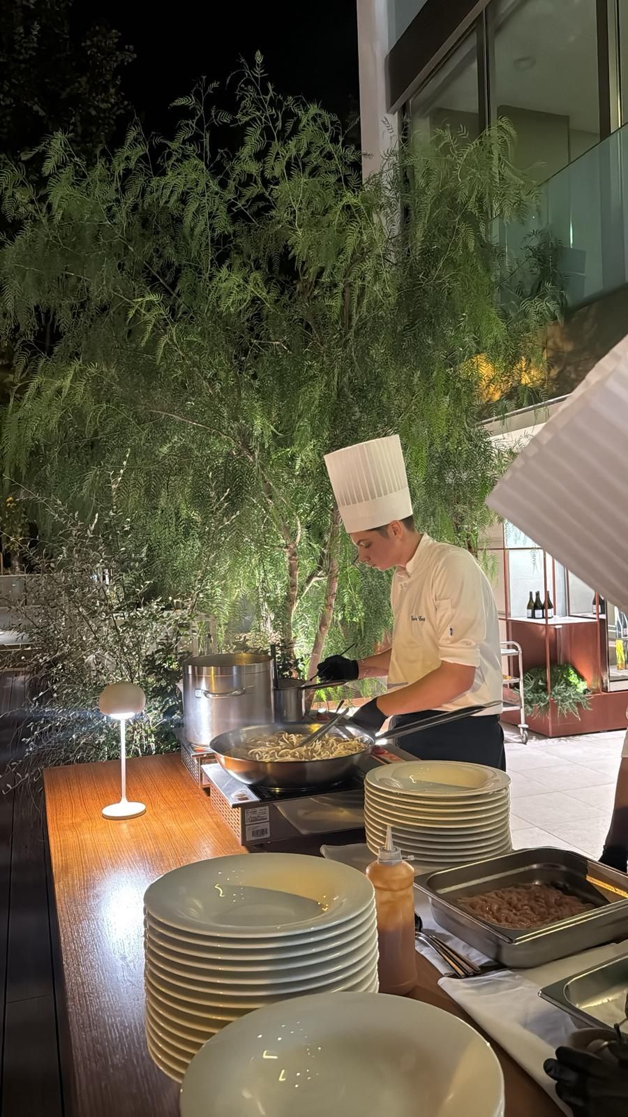 An outdoor dining area with a chef preparing food, surrounded by greenery and a modern building in the background.