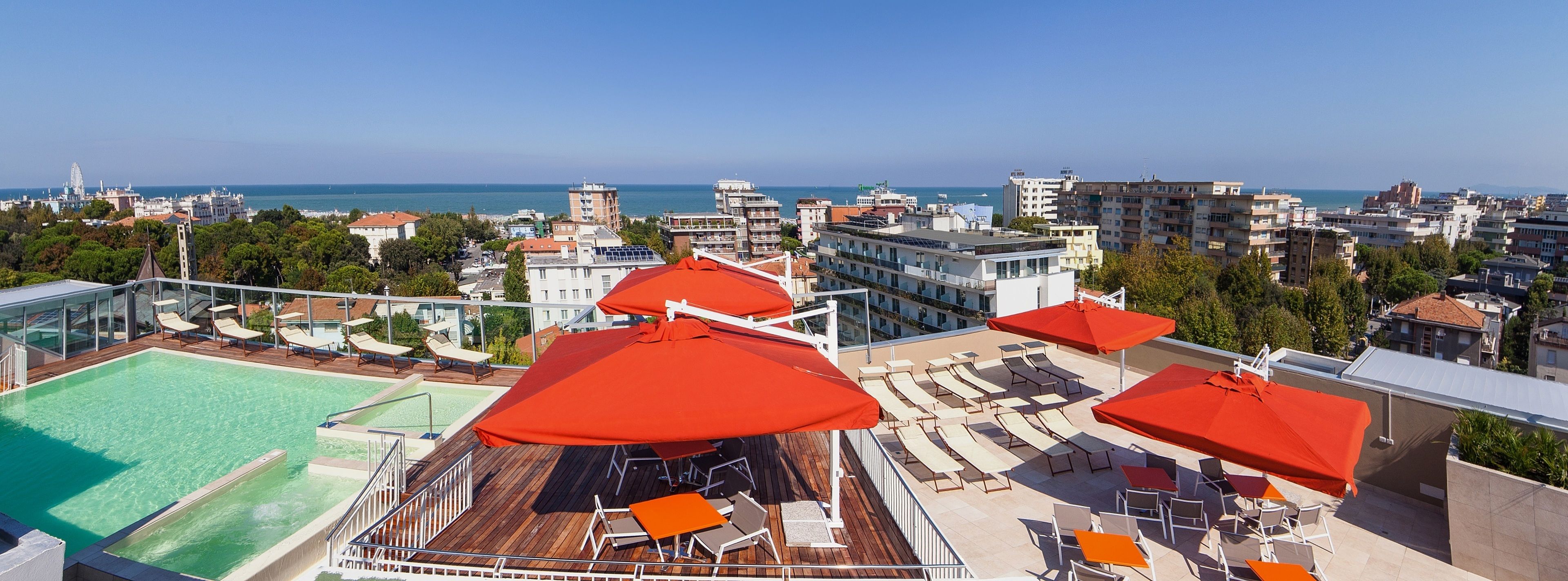 A rooftop pool area with sun loungers and umbrellas, overlooking a cityscape and the sea.