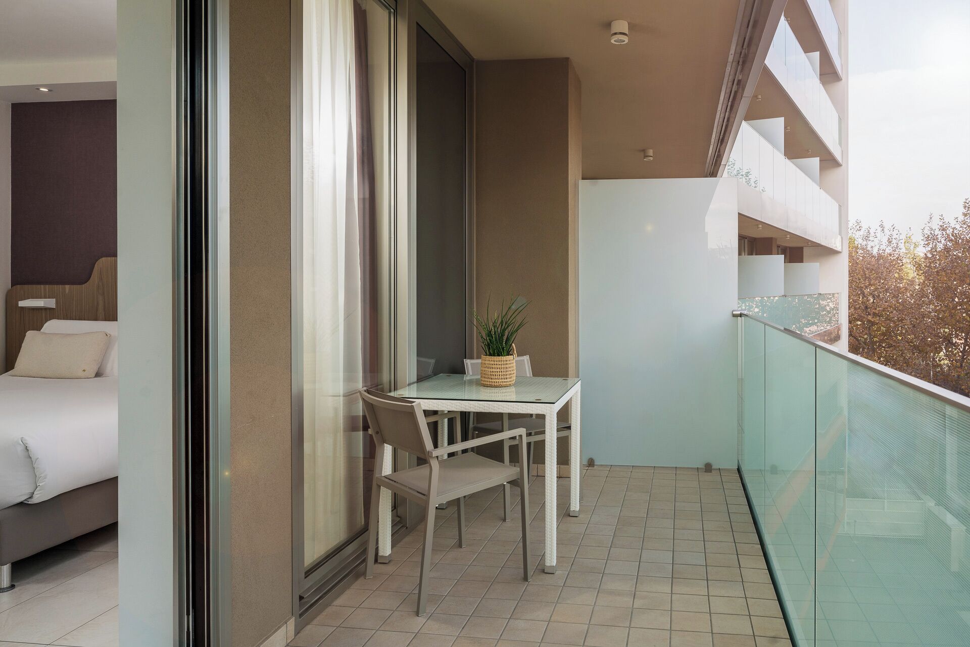 Ein Balkon mit Glastisch und Stühlen, einem Blumentopf und einem Bett im angrenzenden Zimmer.