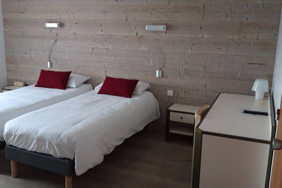 Camera d'albergo con due letti, entrambi con biancheria bianca e cuscini rossi, un comodino con una lampada e una parete con pannello di legno a caposangue.
