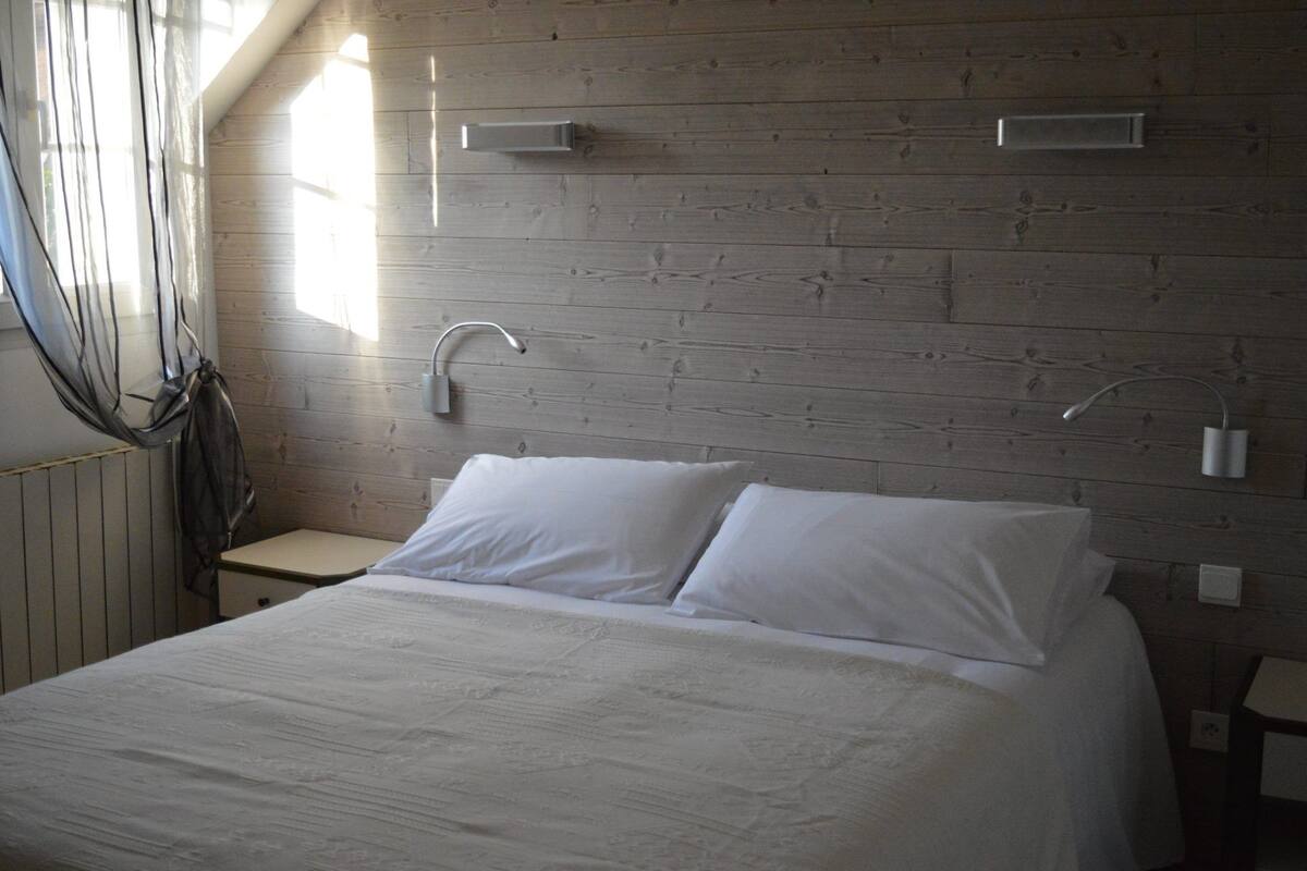 Una camera da letto con testiera in legno, un letto con lenzuola bianche, un comodino e una finestra con tende trasparenti.