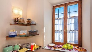 A breakfast setup with various food items and kitchen appliances on a table near a window with curtains.