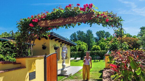 A yellow building with a wooden door, a sign reading "Il Giardino dei Poeti", and a woman in yellow pants standing in front.