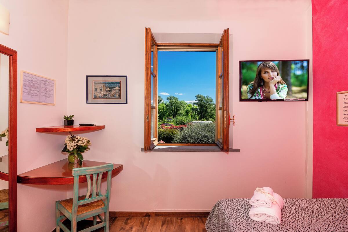 Quarto com escrivaninha de madeira, cadeira, mesinha com um vaso de flores, janela com vista para o verde e cama com toalha.