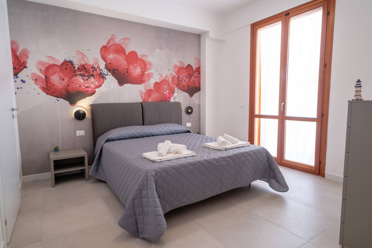 Een slaapkamer met een bed, nachtkastje, muurschildering, raam en kast.