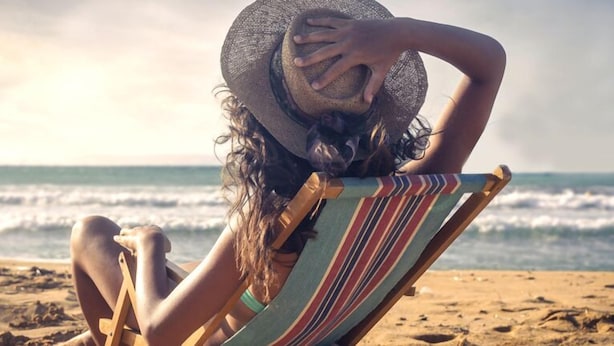 Une personne assise sur une chaise de plage, face à l’océan, portant un chapeau et un maillot de bain.