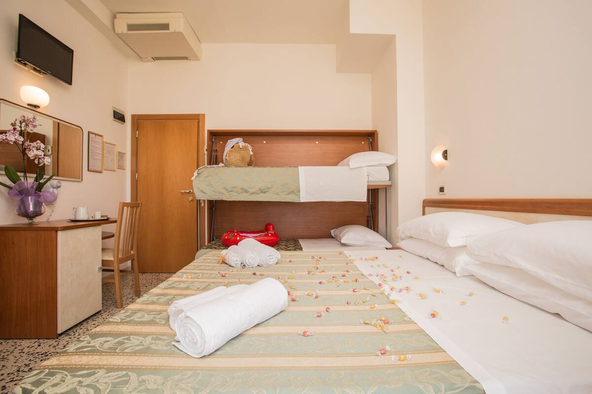 A hotel room with a bunk bed, a desk with a chair, a mirror, and a bedside table with a vase of flowers.