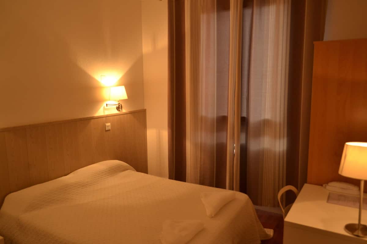 A hotel room with a bed, a nightstand, a lamp, and a view of a curtain and a window.