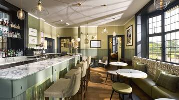 A bar area with a marble countertop, bar stools, and a variety of bottles. There are several tables and chairs arranged for dining, and a sofa with cushions. The room has a green wall and large windows.