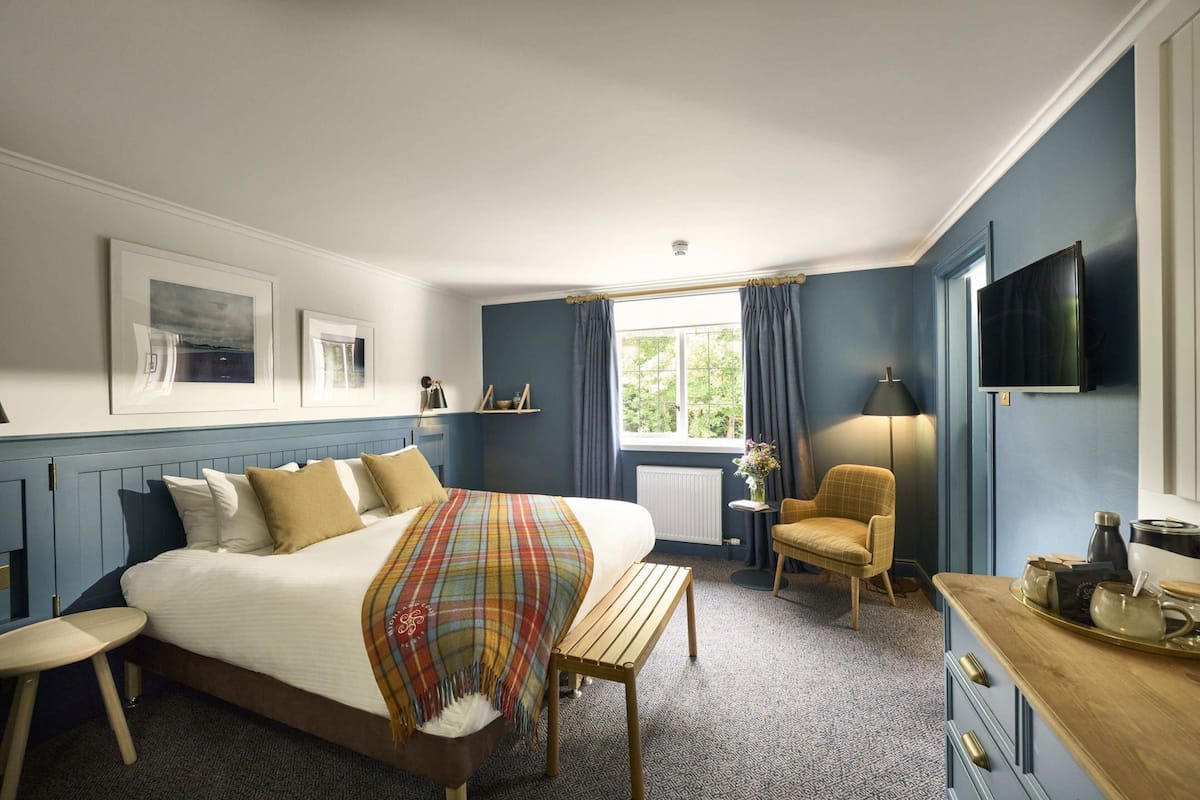 A hotel room with a bed, a chair, a small table, a TV, and a window with curtains.