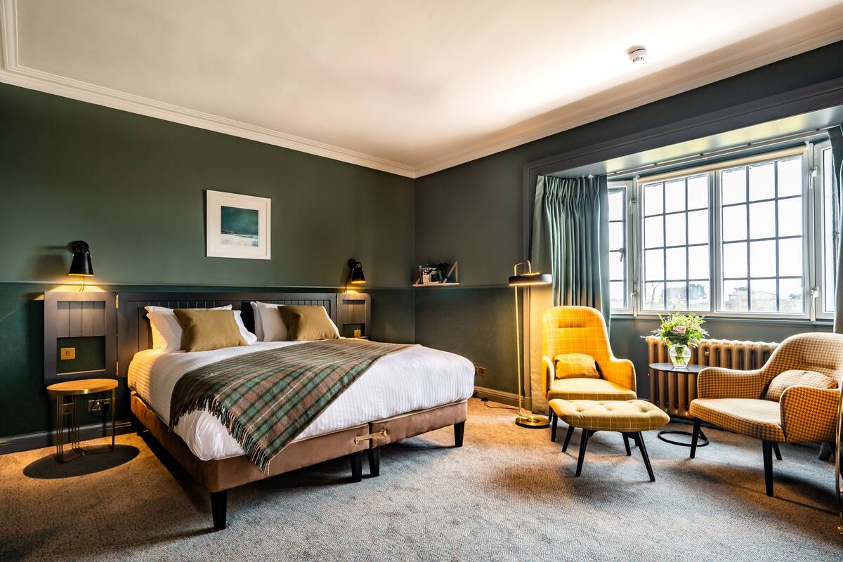 Ein Schlafzimmer mit einem großen Bett, zwei Nachttischlampen, einem Sessel, einem kleinen Tisch und einem Fenster mit Vorhängen.