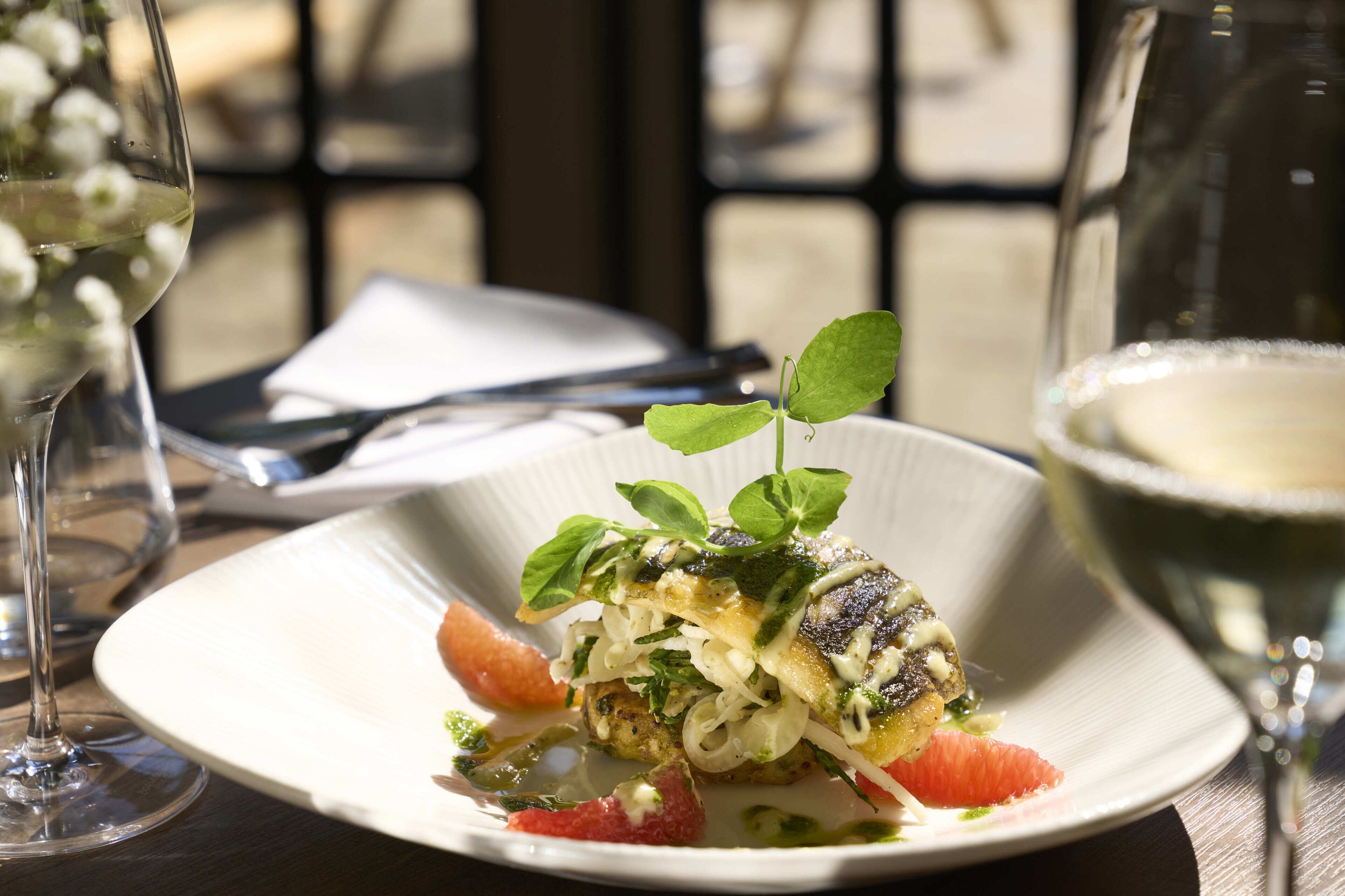 A plate of food with a small green leaf garnish, a glass of white wine, and a fork on a table.