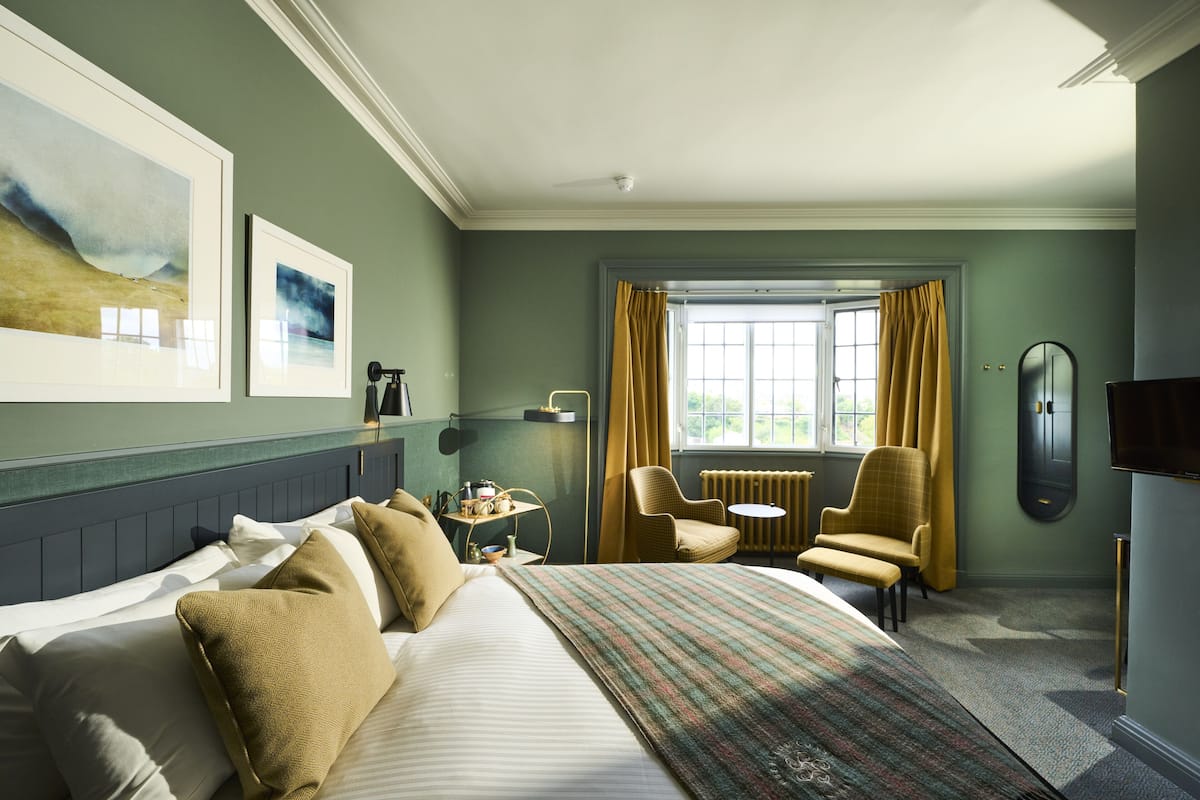 Ein Hotelzimmer mit einem großen Bett, zwei Sesseln, einem kleinen Tisch und einem Fenster mit Vorhängen.